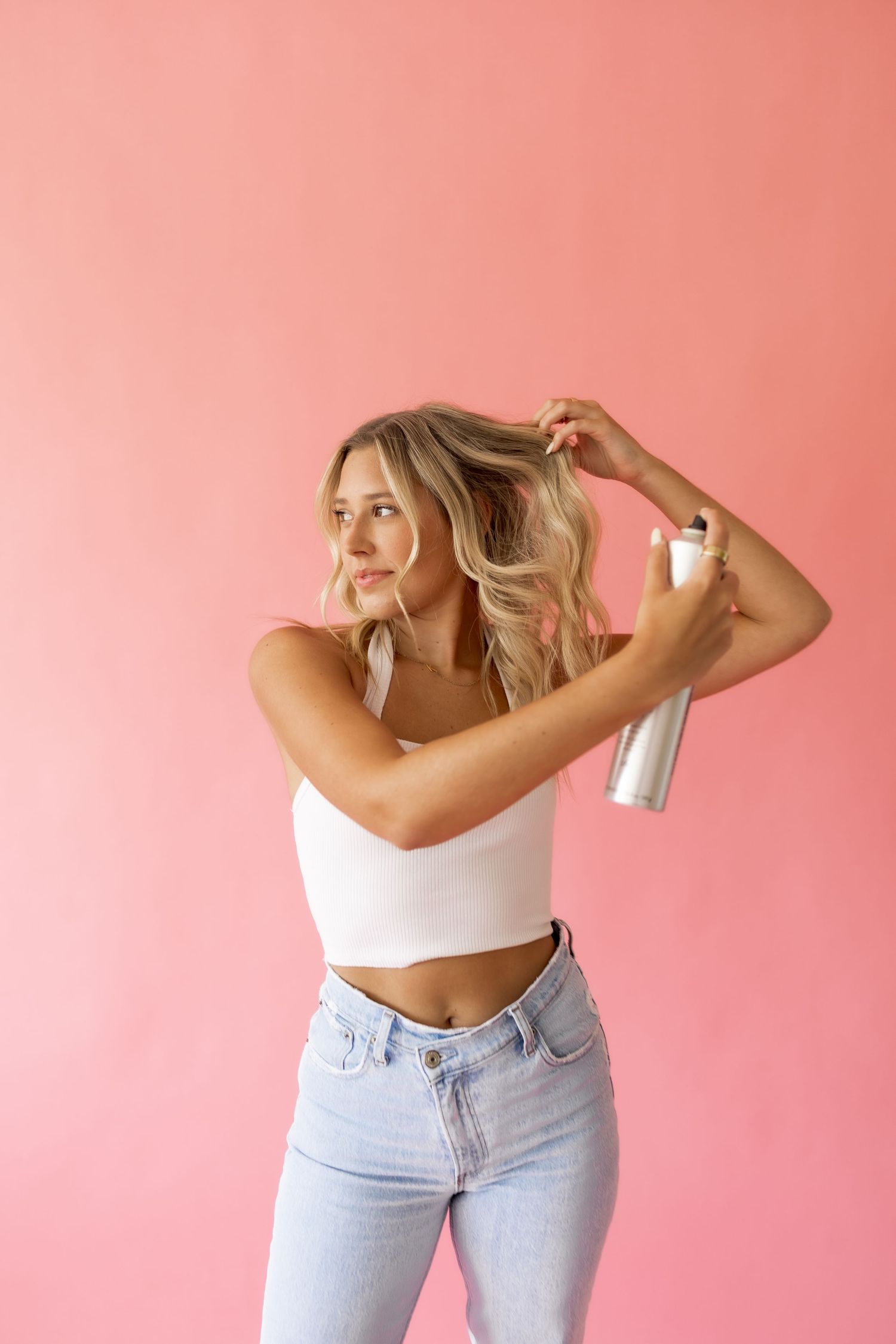 Blonde woman in white crop top and jeans applying hairspray to wavy hair against a pink background.