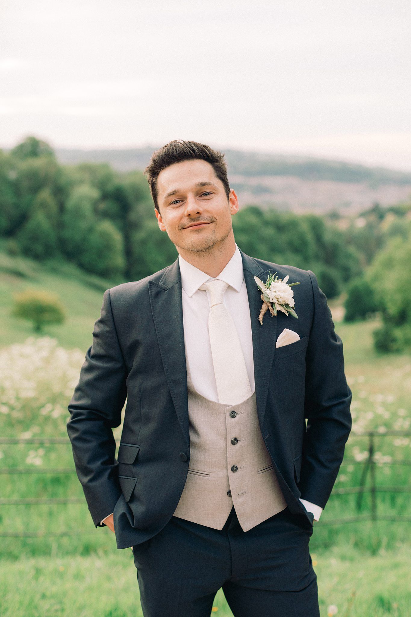 Smiling groom in navy suit with beige vest and floral boutonniere poses outdoors with green hills backdrop.