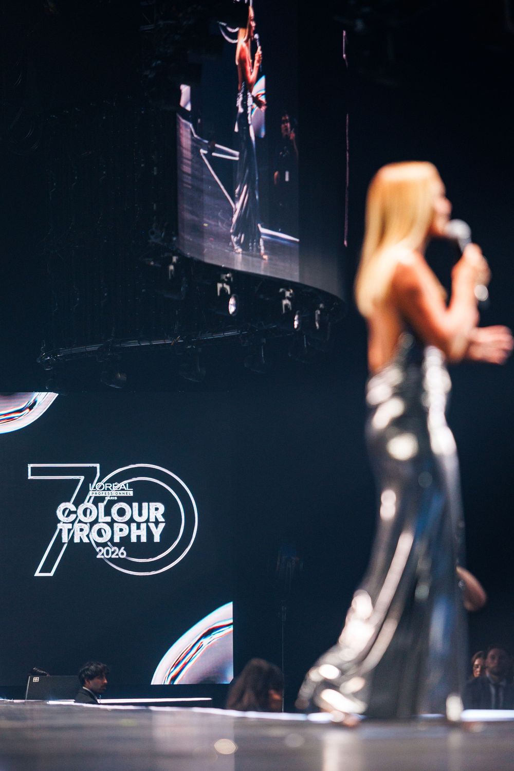 Blonde woman in silver gown presents at L'Oreal Colour Trophy 70th anniversary awards ceremony.