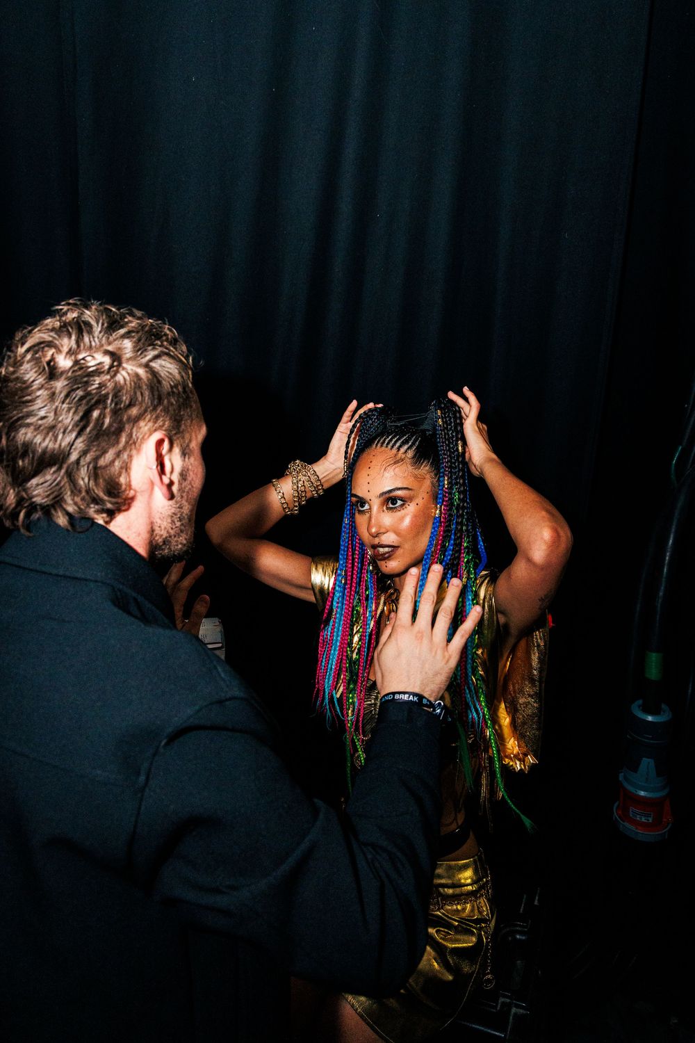 A woman with colorful braids in gold outfit interacts with a man in black jacket against dark curtain backdrop.