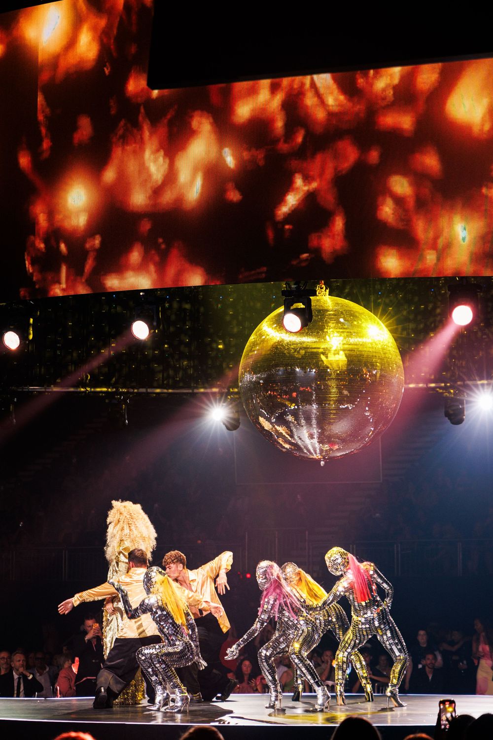 Performers in metallic costumes dance on stage under a golden disco ball with dramatic red lighting.