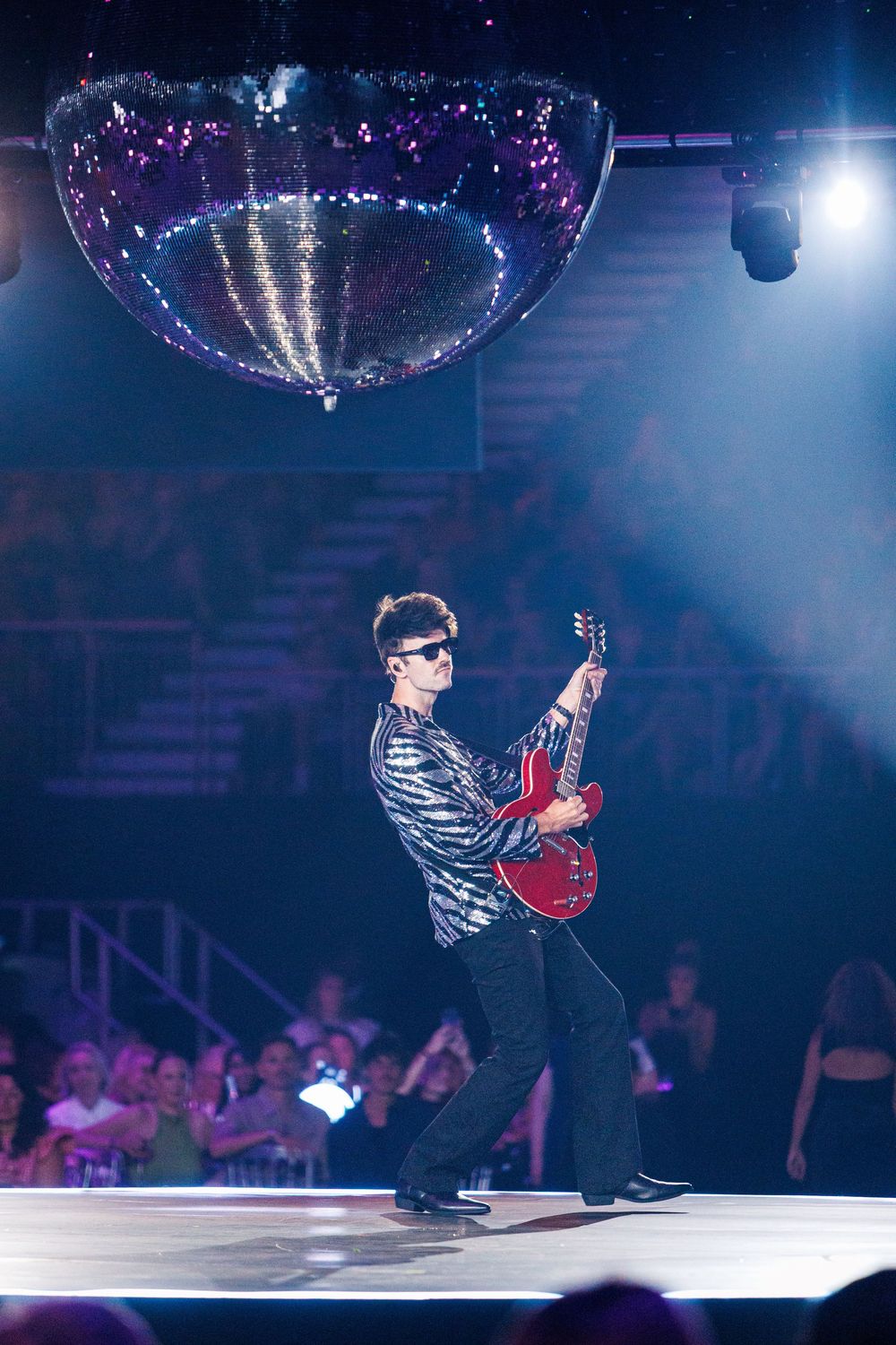 Guitarist performing on stage under a disco ball with crowd in background at a live concert.