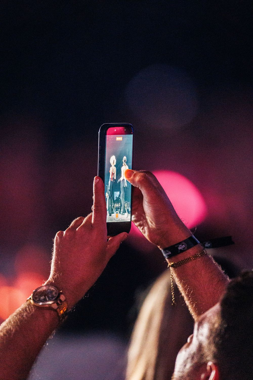 Audience member photographing fashion show performers on smartphone with bokeh lights in background.