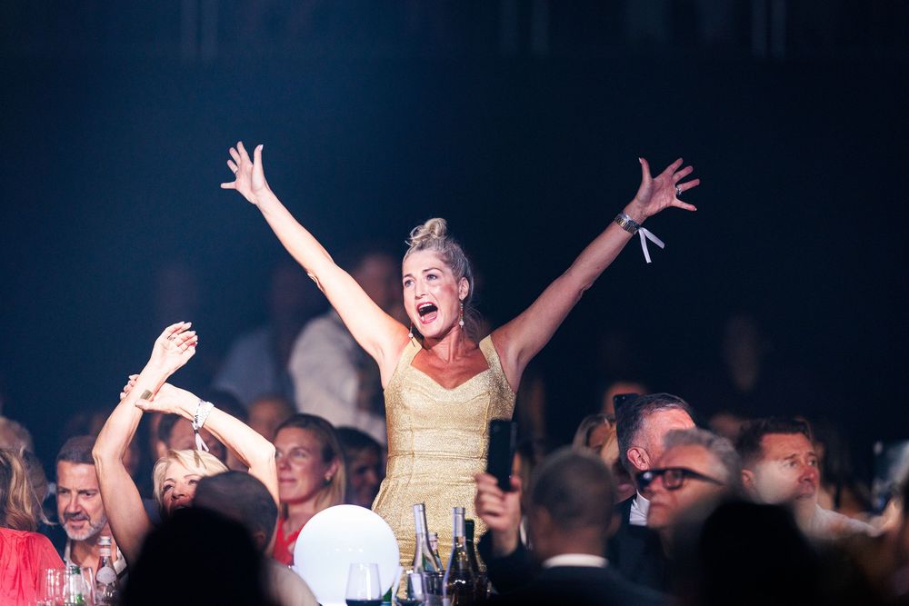 Woman in yellow dress celebrates with arms raised at a lively formal event or gala dinner party.