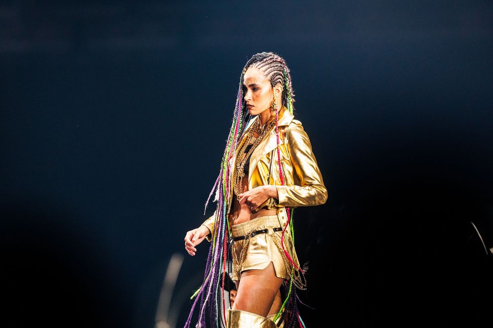 Female performer in gold outfit with colorful braids on stage under dramatic lighting.