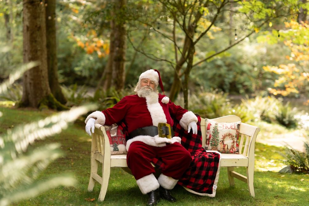 Santa Claus sitting relaxed on a white bench outdoors in a lush green garden wearing his classic red suit.