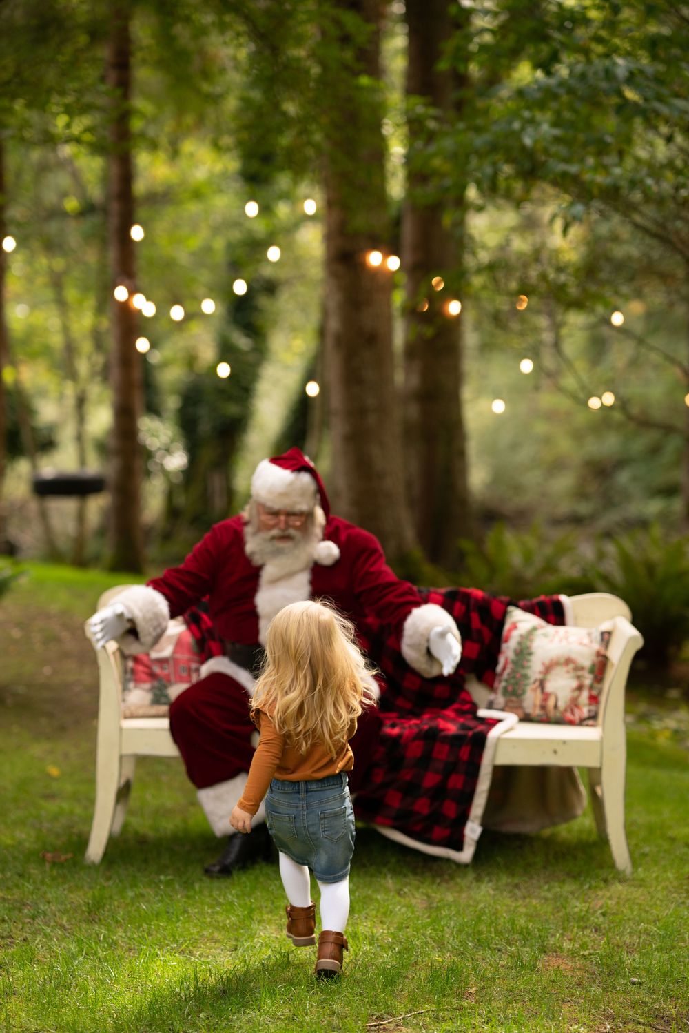 Young child runs across green grass toward Santa Claus seated outdoors among trees with string lights.