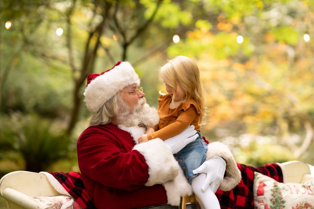 A child sits on Santa Claus's lap in a festive outdoor setting with autumn foliage in the background.