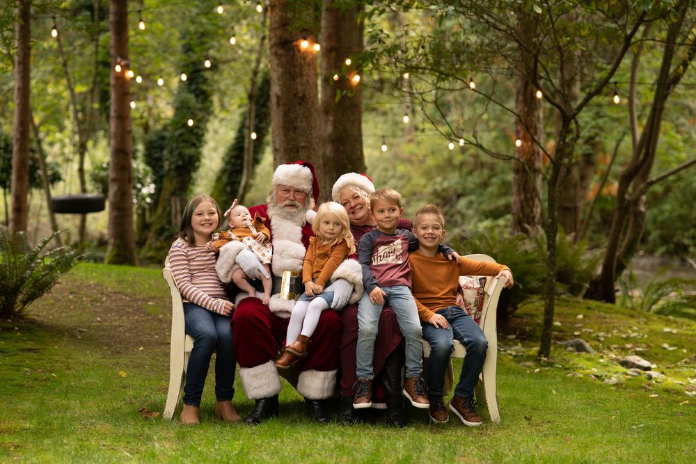 Santa Claus poses with a group of five children on a bench in a lush outdoor forest setting.