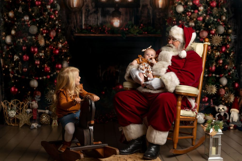 Santa Claus sits in a rocking chair reading to children by a decorated Christmas tree and fireplace.