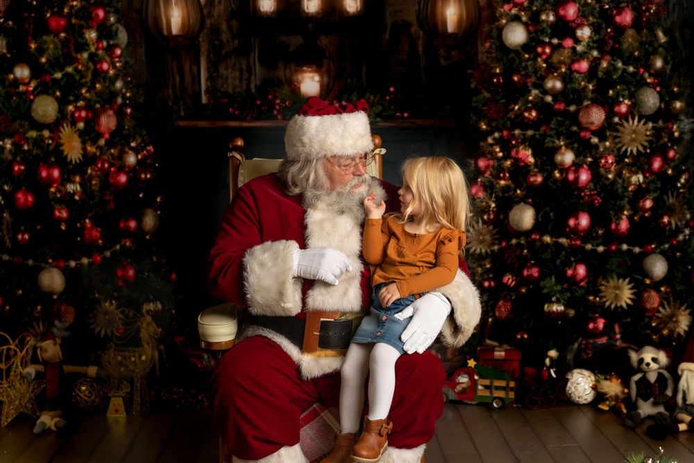 A young child sits on Santa Claus's lap whispering in his ear beside decorated Christmas trees.
