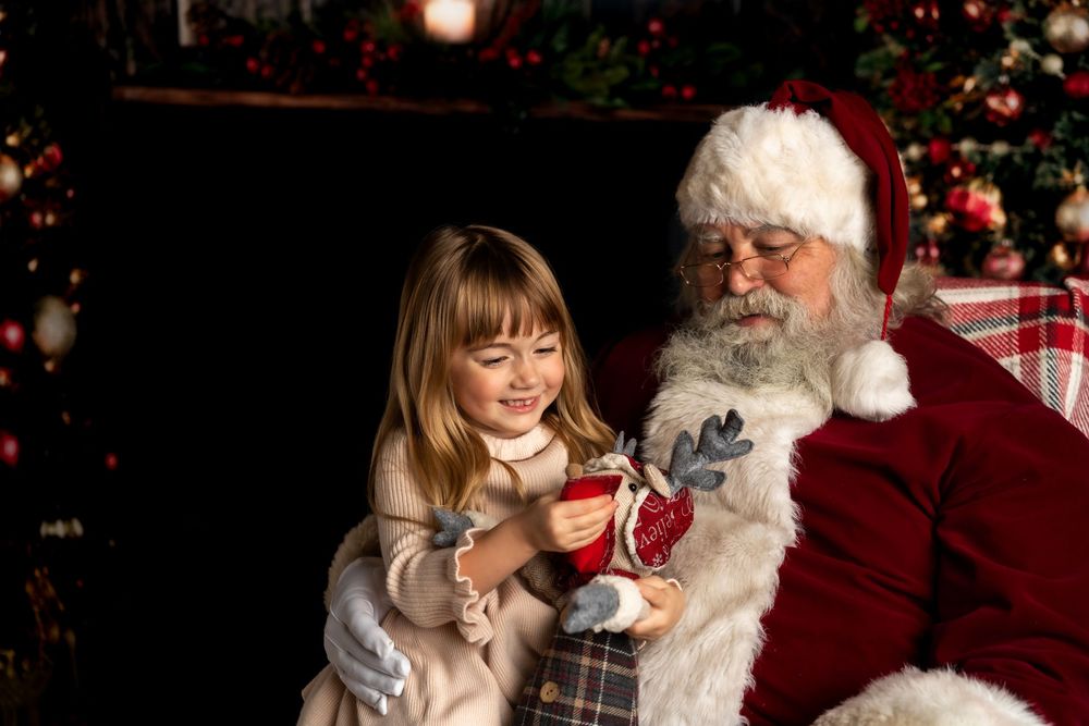 A young girl sits on Santa Claus's lap opening a Christmas gift near a decorated tree.