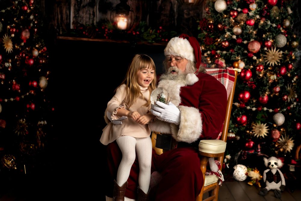 A young girl sits on Santa Claus's lap examining a snow globe in a festive Christmas setting.