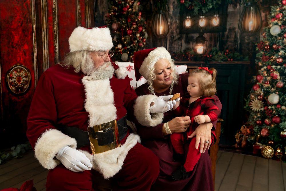 Santa and Mrs. Claus reading a list with a young girl beside a decorated Christmas tree indoors.