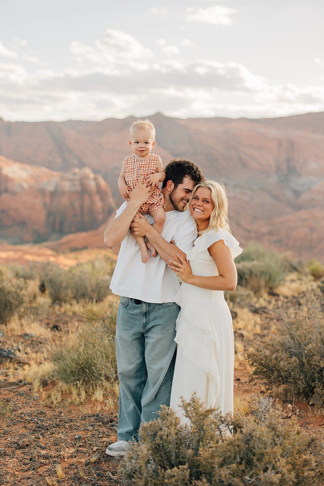 Happy family with baby posing in red rock desert landscape at golden hour, man in jeans and woman in white dress.