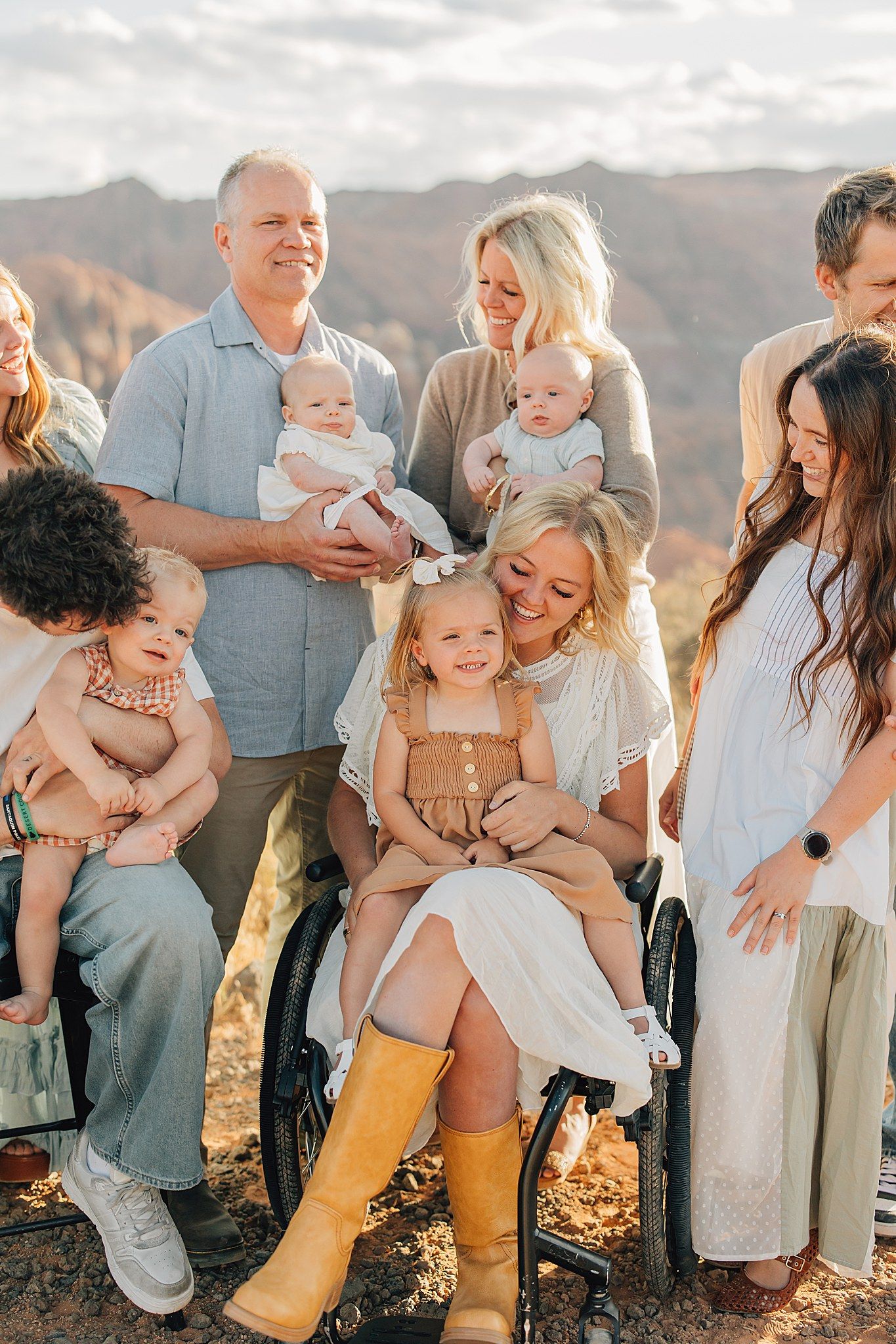 Large family group photo outdoors with babies, toddlers, and adults smiling, woman in wheelchair holding young girl.