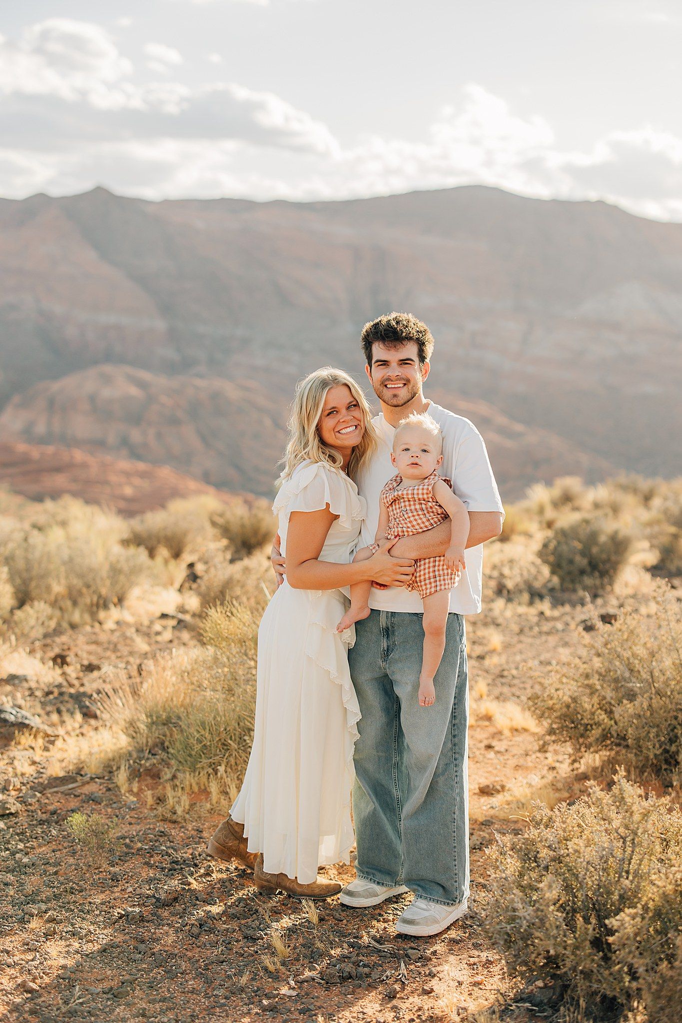 A young family smiles outdoors, woman in white dress, man in jeans holding a baby, with red rock mountains behind.
