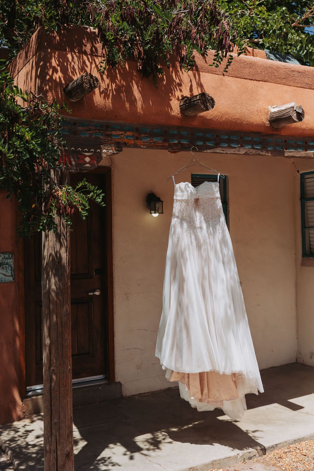 White wedding dress hanging on a wooden beam outside a rustic adobe building with warm terracotta walls and lush greenery.