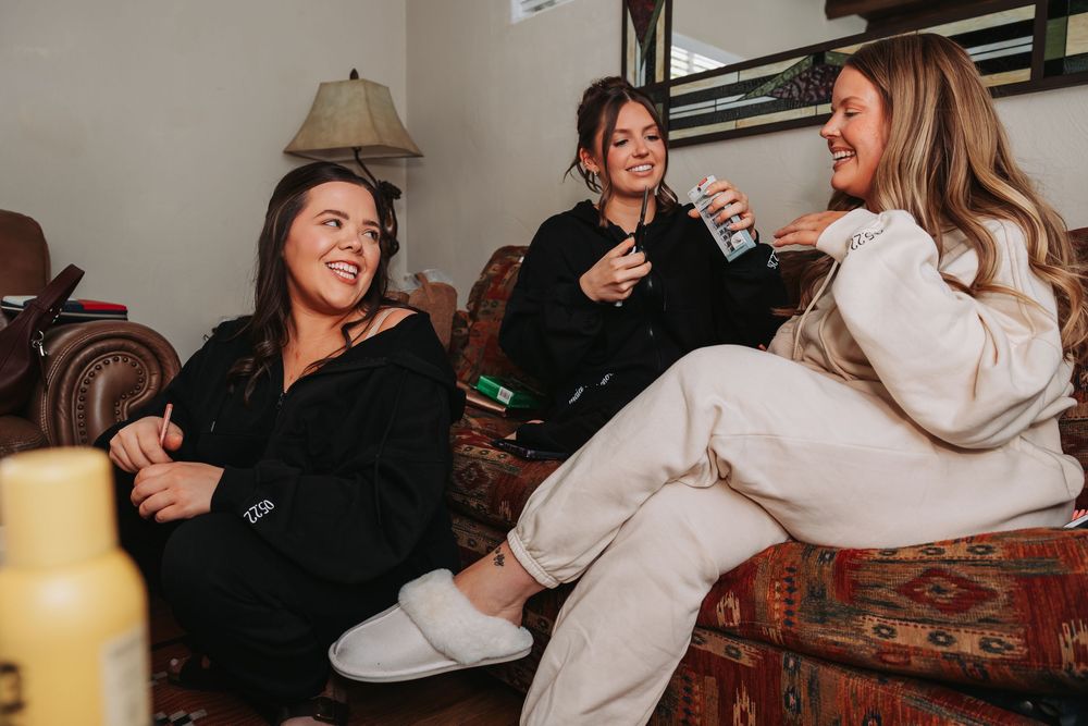 Three women laughing together on a couch in robes and slippers before a wedding.