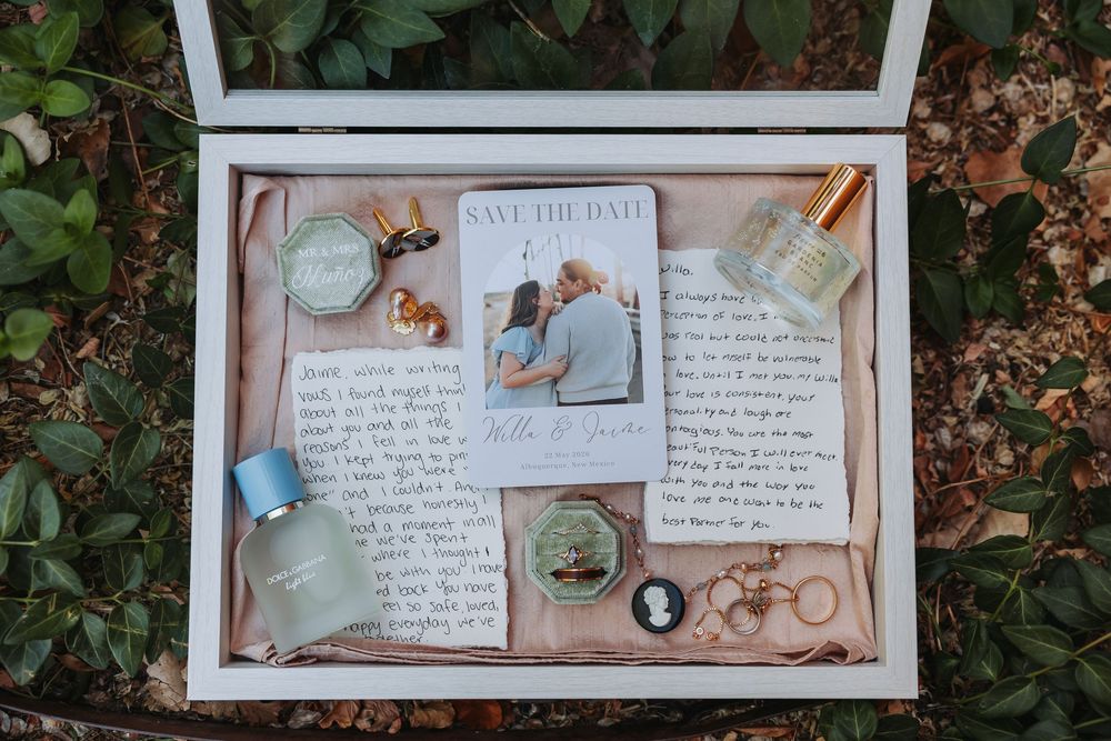 A wedding save the date card displayed in a shadow box with perfume bottles, rings, and love letters on a floral background.
