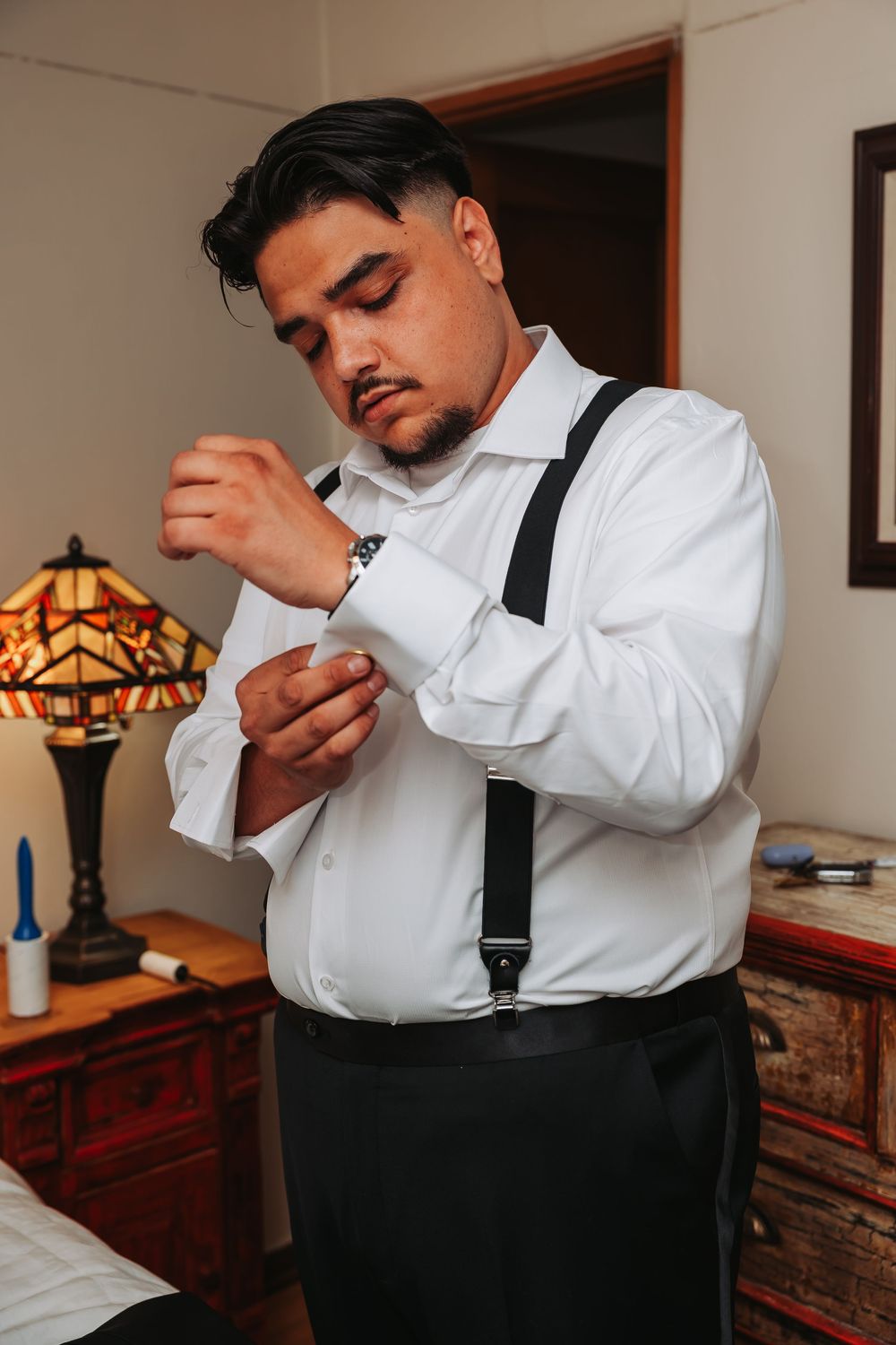 A man in white dress shirt and suspenders fastening his cufflinks while getting ready for a formal event.