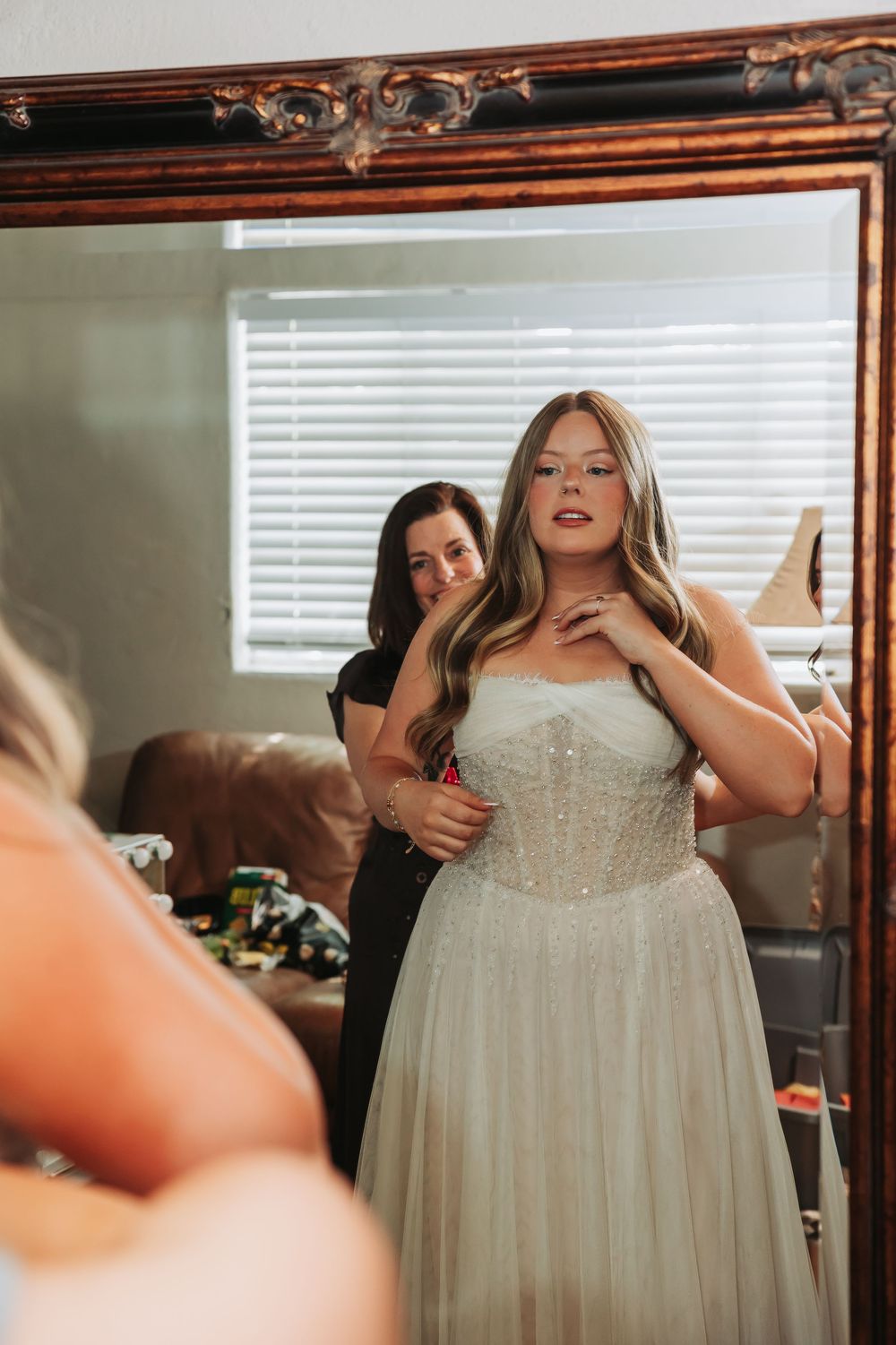 Bride in strapless white wedding gown being helped by bridesmaid, reflected in ornate mirror while getting ready.