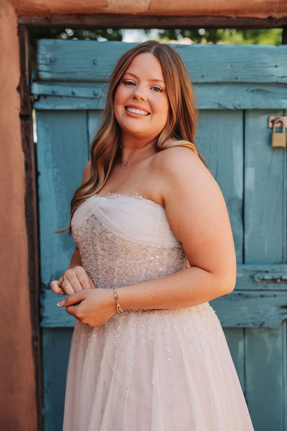 Smiling bride in strapless beaded gown poses before rustic turquoise door in warm golden light.