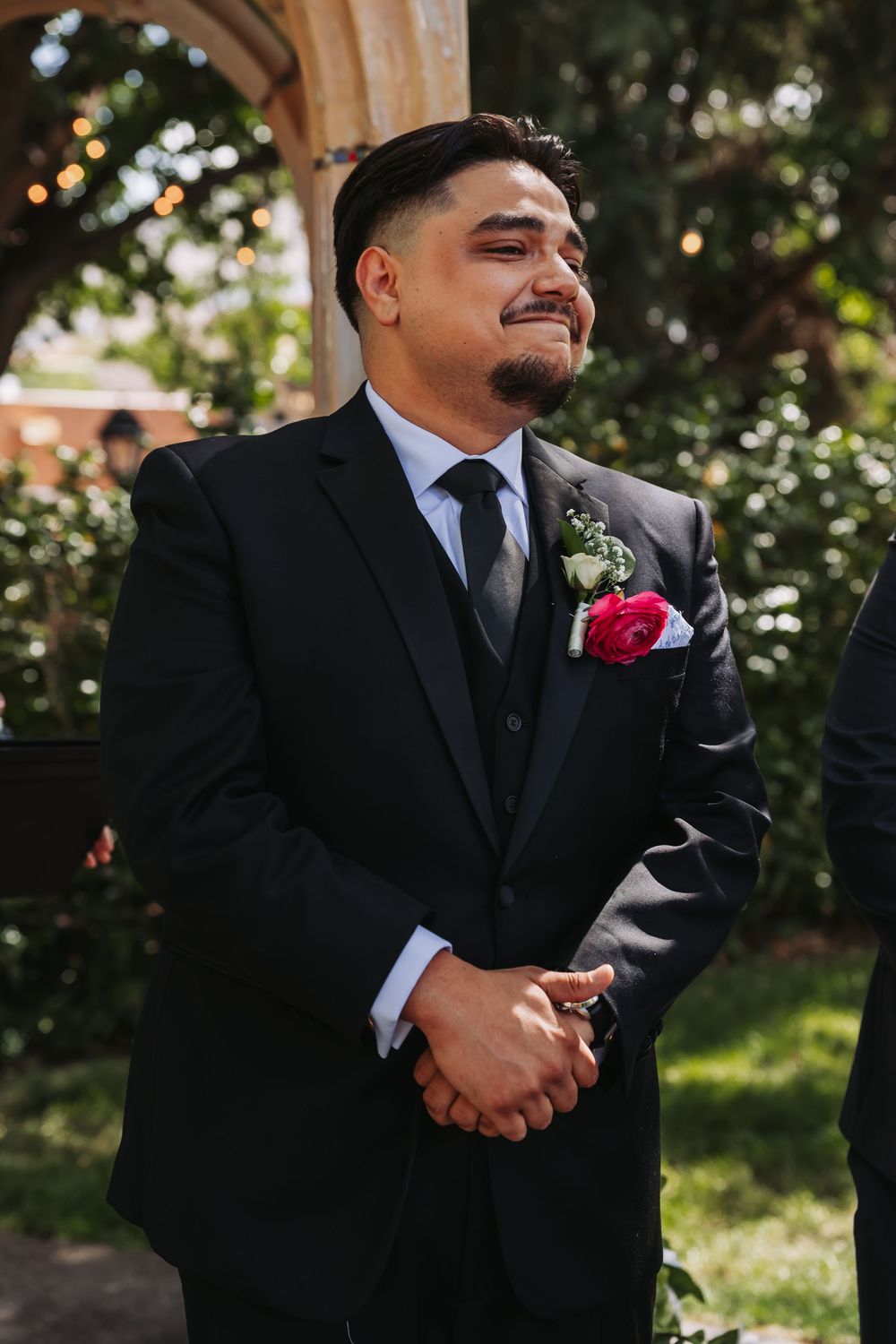 Groom in black suit with red boutonniere smiling emotionally during outdoor wedding ceremony.