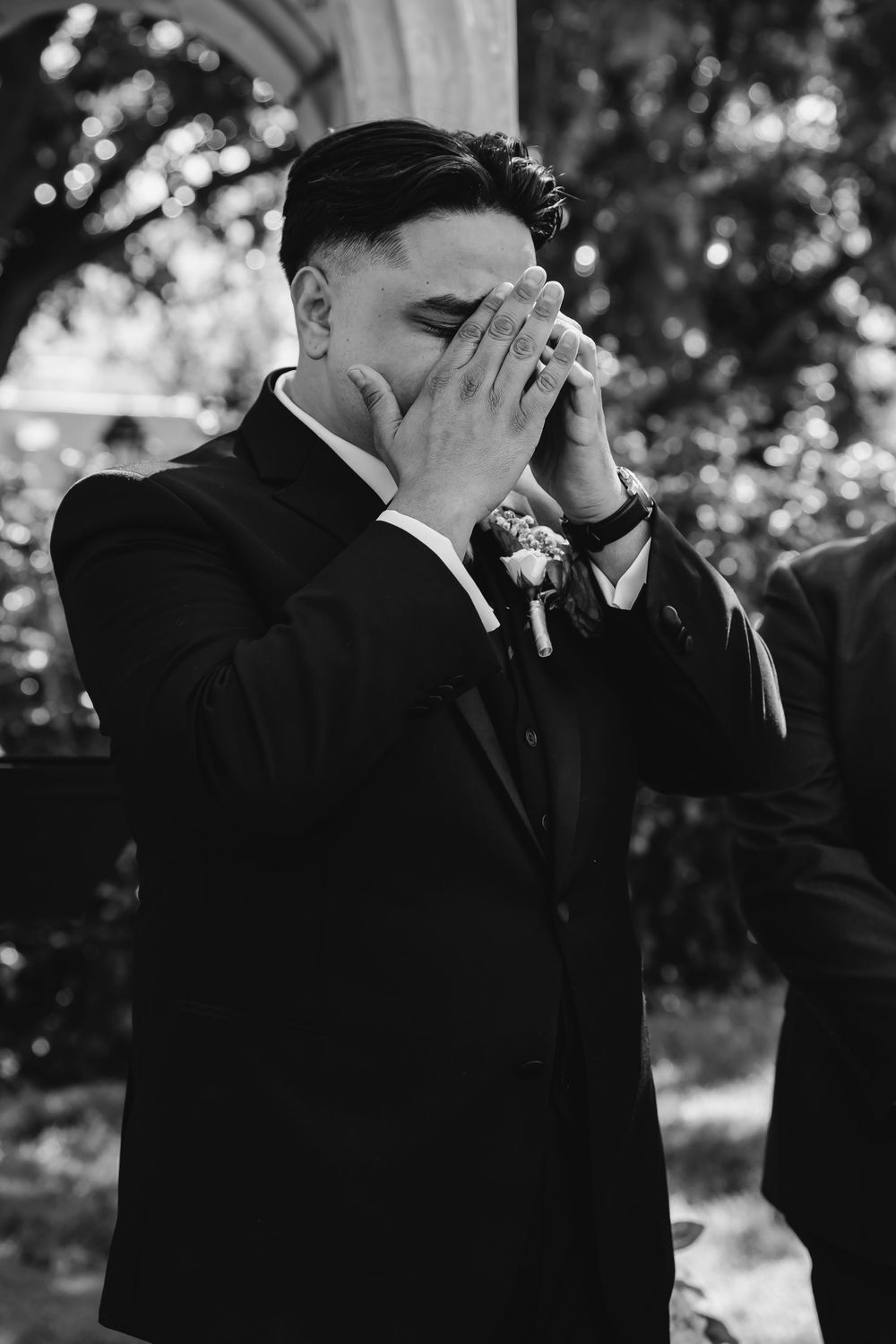 Emotional groom in black tuxedo covering his face with hands during wedding ceremony, black and white photo.