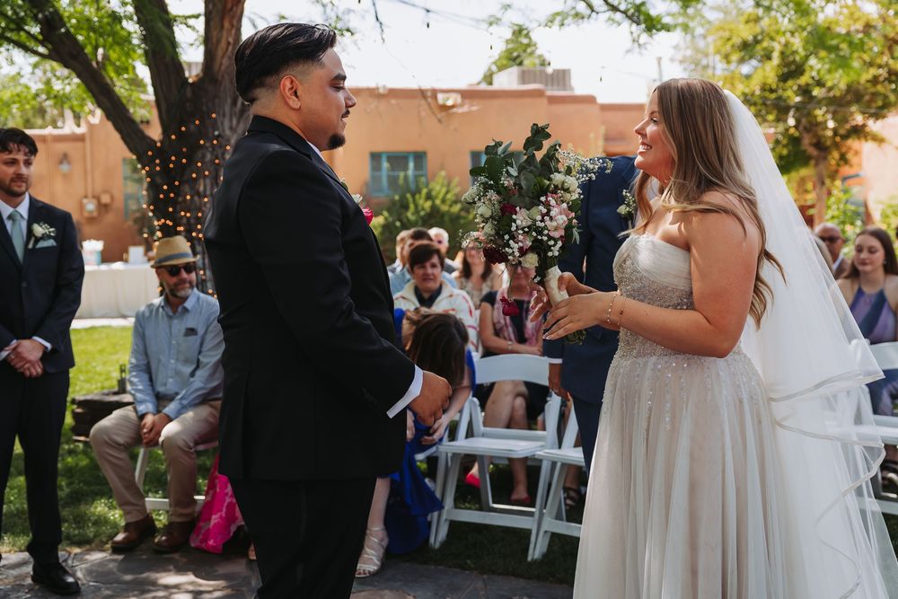 Bride and groom exchange vows outdoors at a sunny wedding ceremony with guests seated in white chairs.