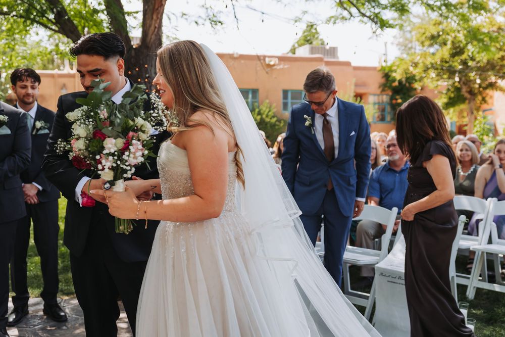 Bride in white gown and veil walks down outdoor aisle with groom in navy suit during wedding ceremony.