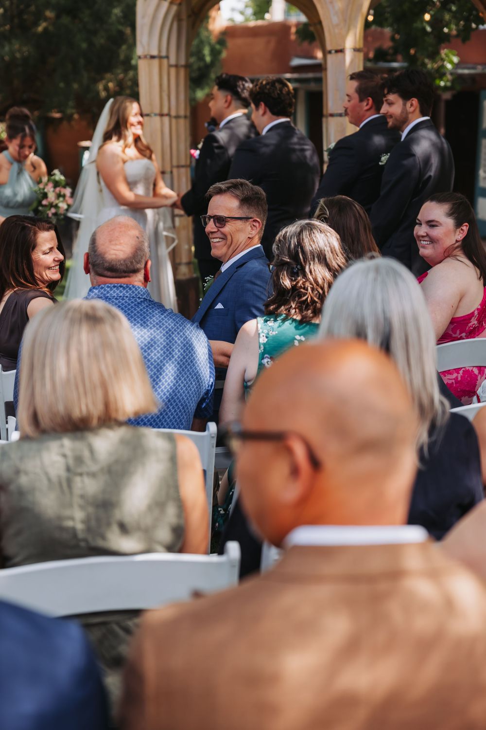 Guests smiling and laughing during an outdoor wedding ceremony seated in white chairs.