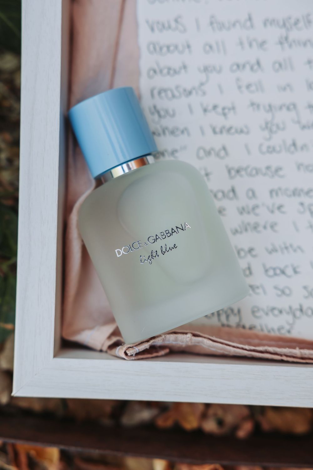 Dolce & Gabbana Light Blue perfume bottle with blue cap resting on a handwritten love letter in a white frame.