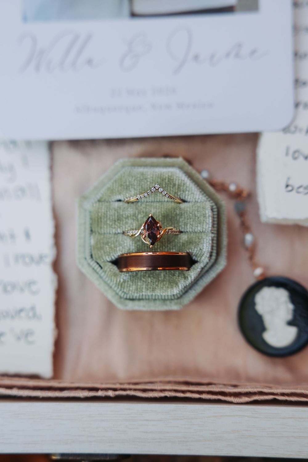 Rose gold engagement ring with purple gemstone in green velvet ring box, wedding invitation and cameo brooch nearby.