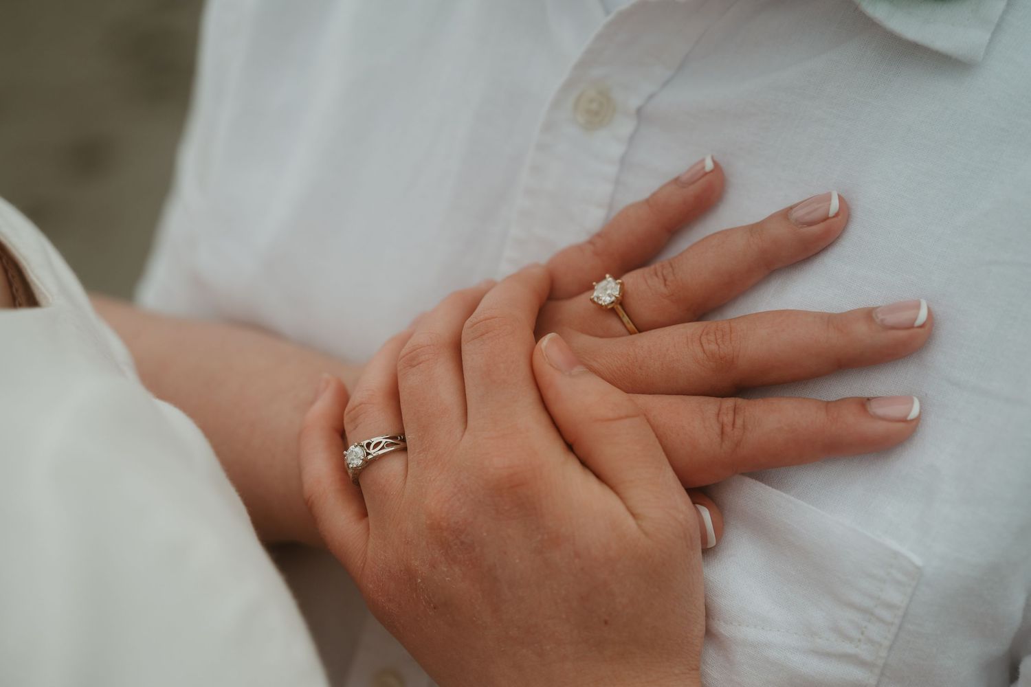 Close-up of two hands with elegant engagement rings, one featuring a solitaire diamond, resting on a white shirt.