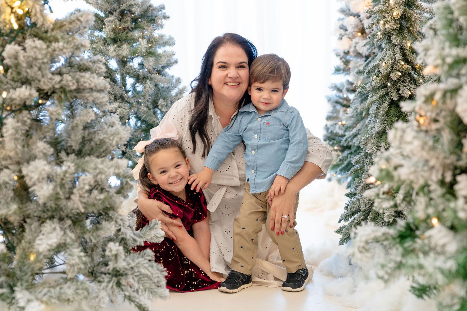 A mother poses with two young children among snow-flocked Christmas trees in a festive holiday photo session.