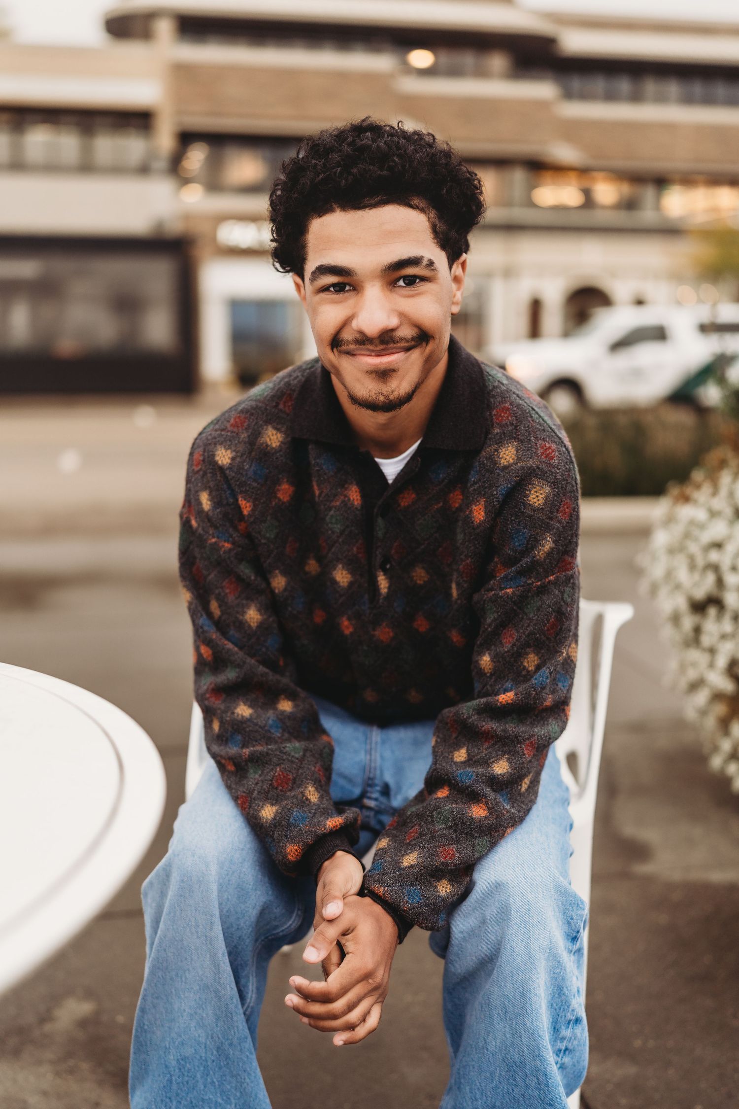 Young man with curly hair smiling, wearing a patterned sweater and jeans, sitting outdoors in an urban setting.