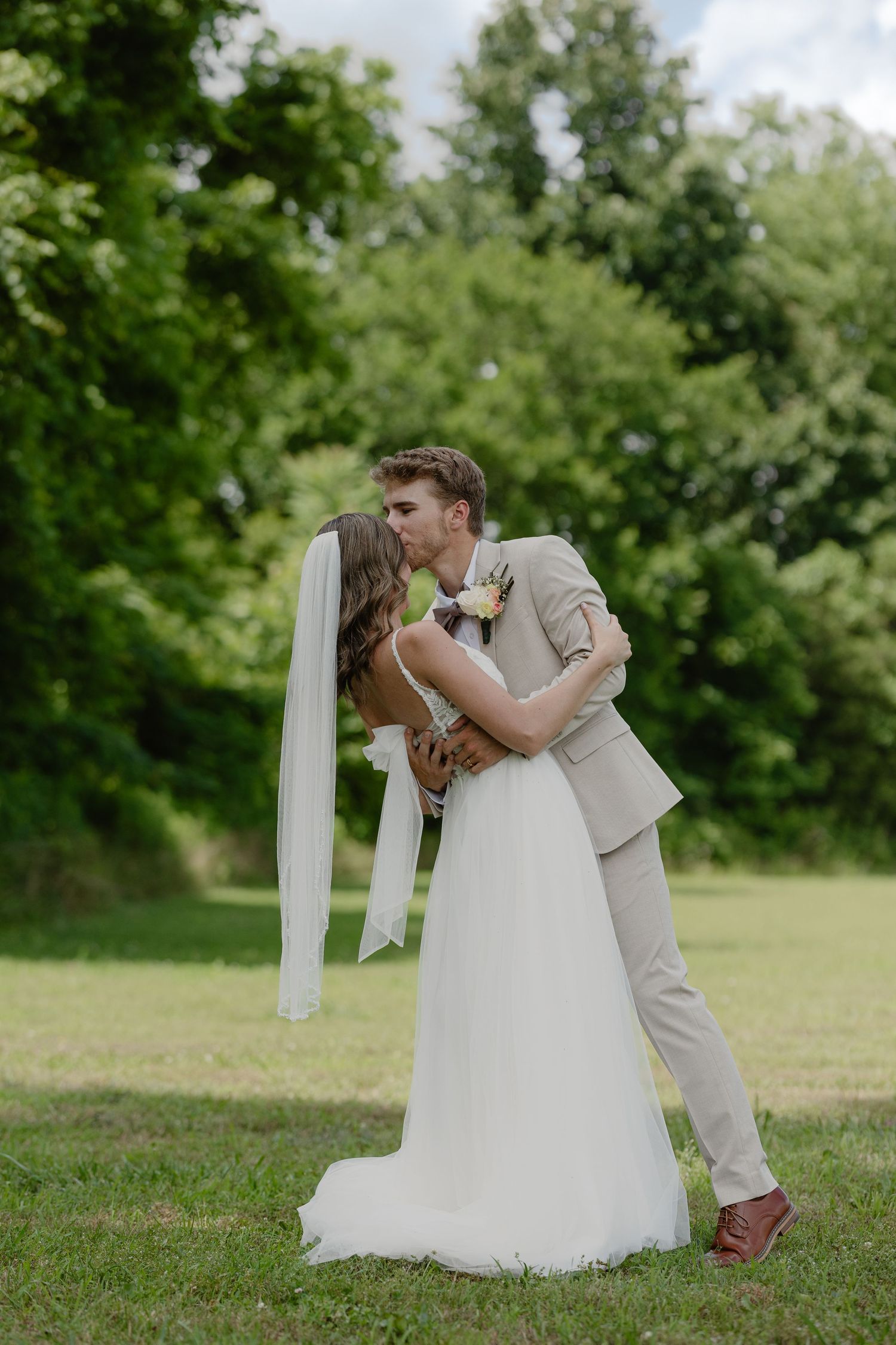 Bride and groom share a romantic kiss outdoors on green grass with lush trees in the background.
