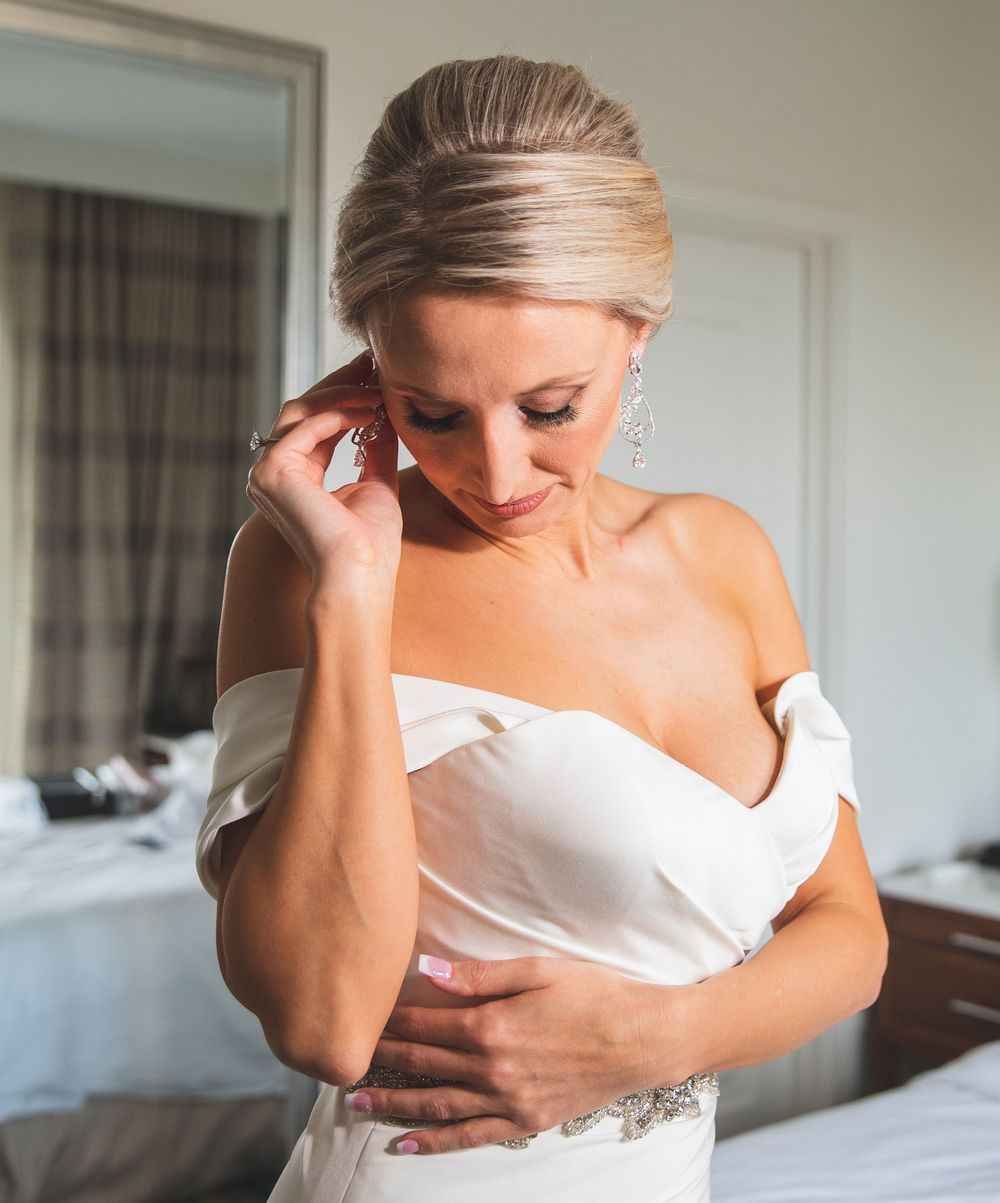 Bride in white off-shoulder gown adjusting earring, elegant updo and crystal jewelry, soft natural light.