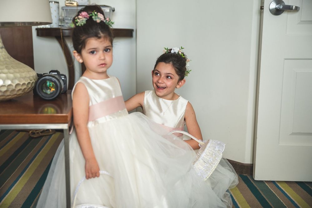 Two young flower girls in white tulle dresses with floral crowns pose together in a bright room.