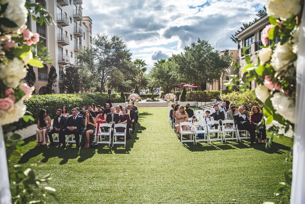 Outdoor wedding ceremony on green lawn with white chairs, floral arch, and hotel backdrop under dramatic cloudy sky.