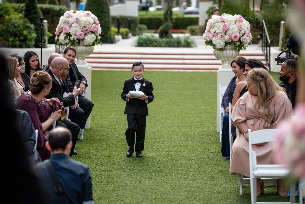 Young ring bearer in black tuxedo walks down outdoor wedding aisle flanked by seated guests and pink floral arrangements.