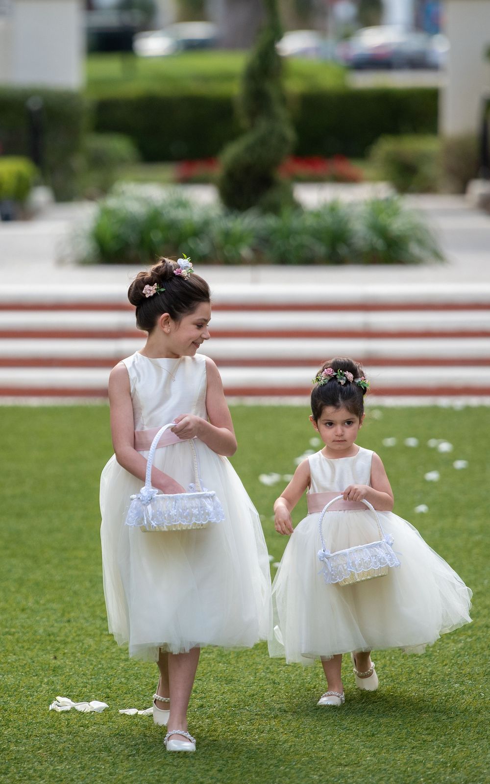 Two flower girls in white tulle dresses carrying baskets walk across green lawn at outdoor wedding ceremony.
