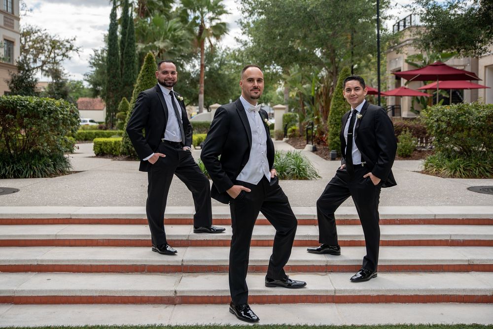 Three groomsmen in black tuxedos pose playfully on outdoor steps at a wedding venue surrounded by palm trees.