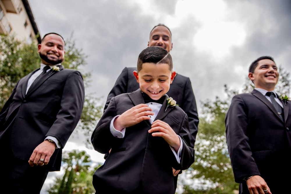 Young ring bearer in black suit adjusts boutonniere, flanked by smiling groomsmen outdoors on a cloudy wedding day.