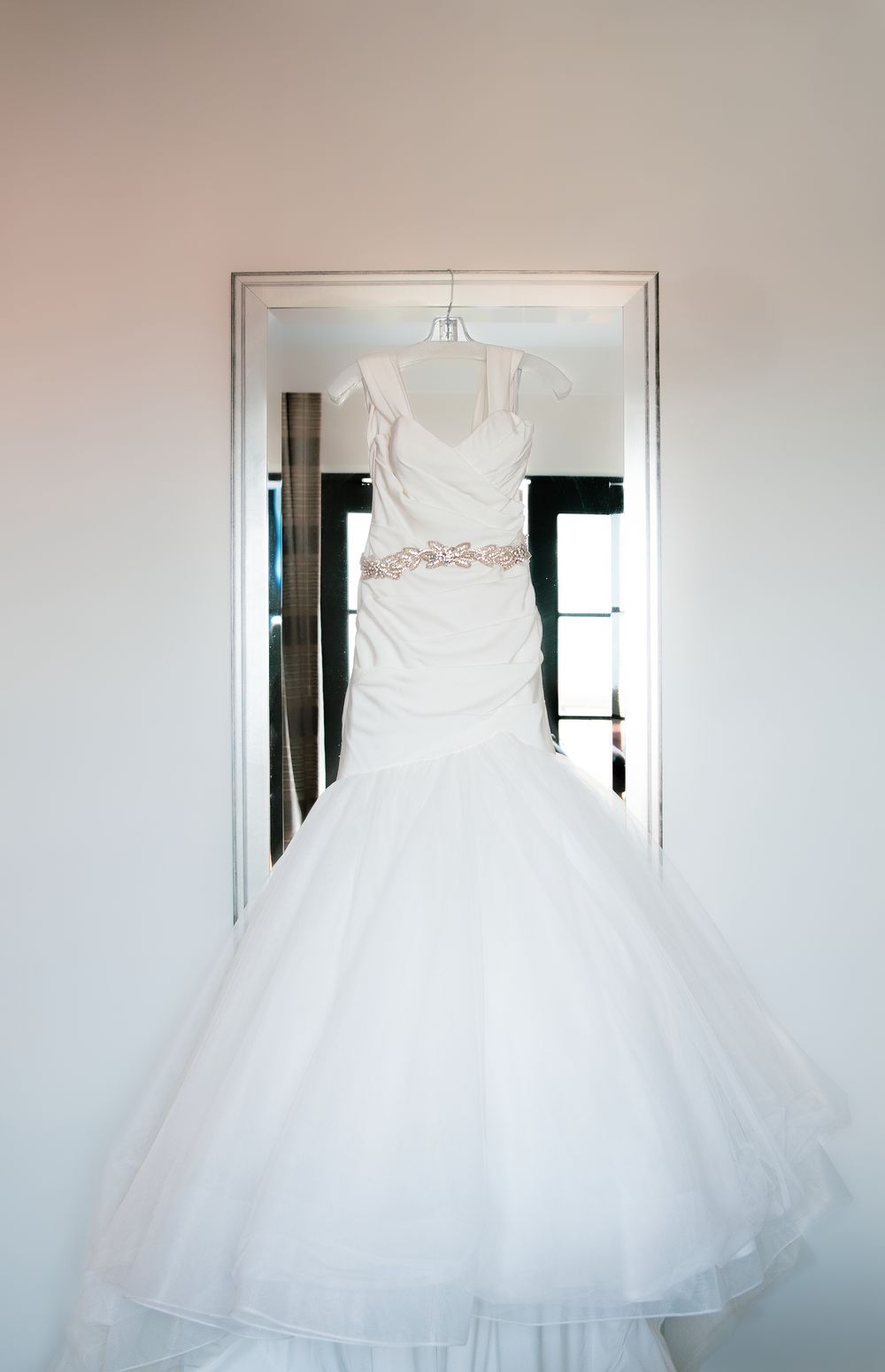 Elegant white mermaid wedding dress with pink sash hanging in a doorway on a hanger.