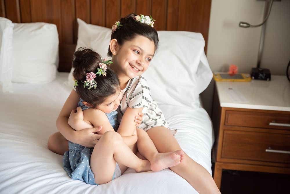 Two young girls with flower crowns hugging and smiling on a white bed in a bright hotel room.