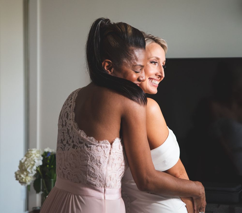 Two women in elegant dresses embrace warmly, one in white bridal gown and one in pink lace dress.