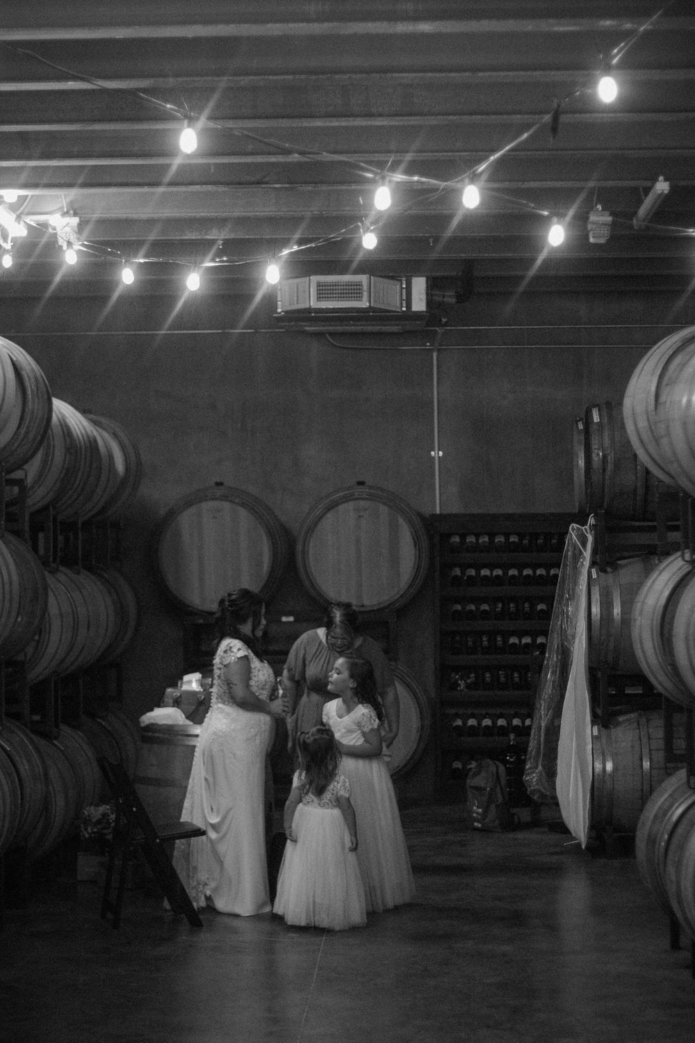 Black and white photo of a wedding party among wine barrels in a winery, with string lights overhead.