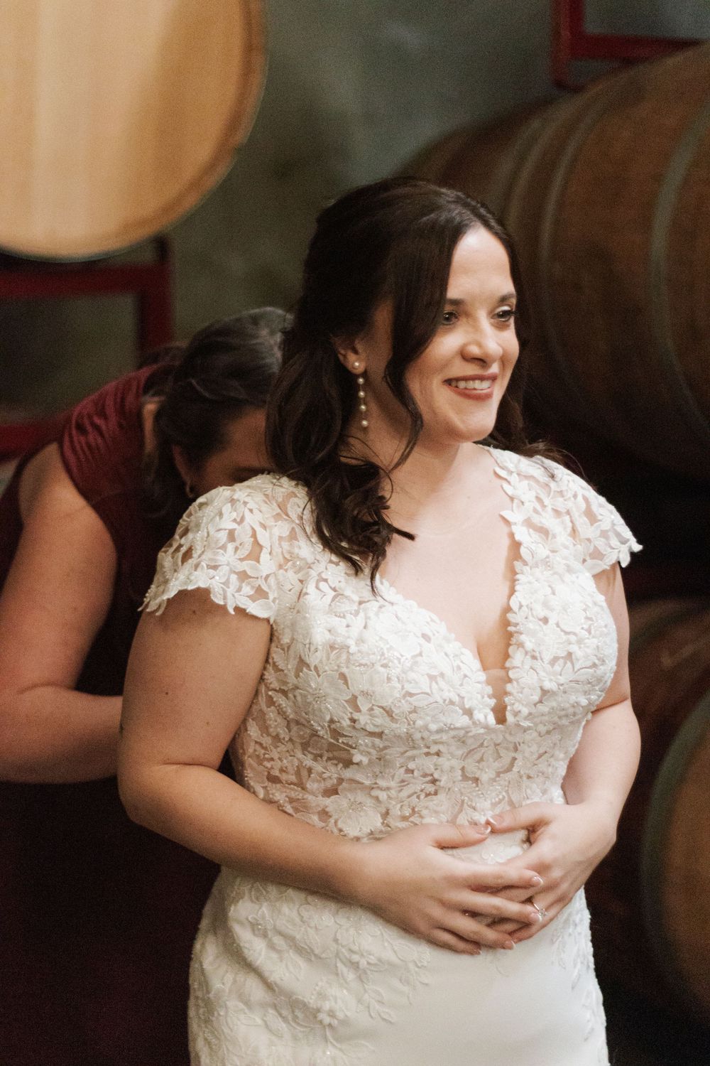 Bride in lace cap-sleeve wedding gown smiles as bridesmaid in burgundy dress helps her prepare near wine barrels.